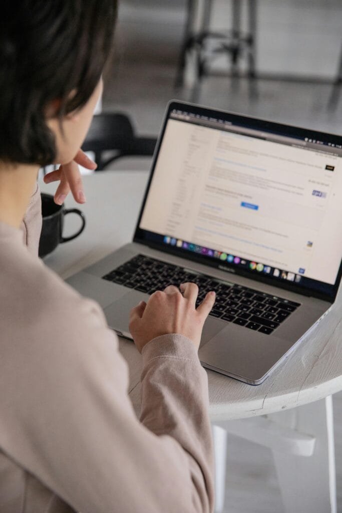 Back view of crop anonymous female remote employee typing on portable computer while watching website on screen and sitting at plastic table with cup of hot drink in apartment